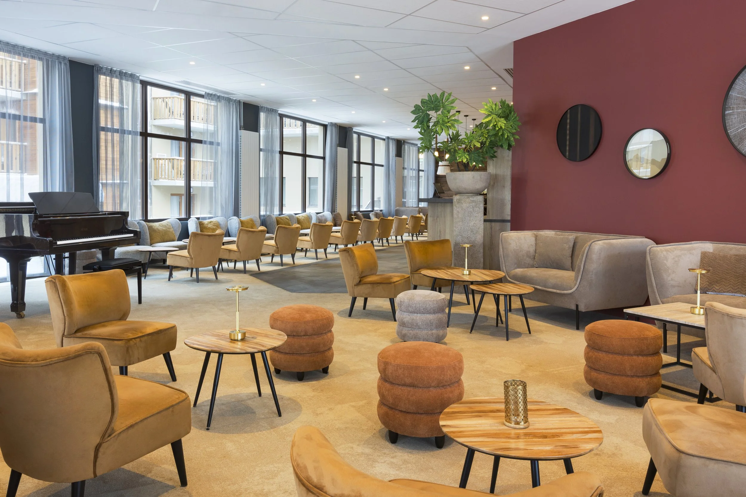 Salon d'hôtel avec des tables basses en bois, des chaises beige, des canapés gris, un piano, une plante verte, un mur couleur bordeaux avec des miroirs accrochés, et plusieurs baies vitrées.