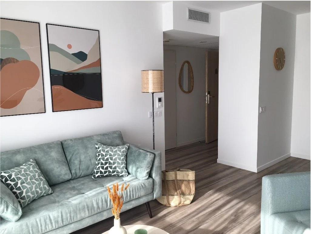 Mur blanc avec des tableaux géométriques orange blanc et bleu. Une lampe, un canapé bleu clair ainsi qu'un miroir et une horloge au fond.