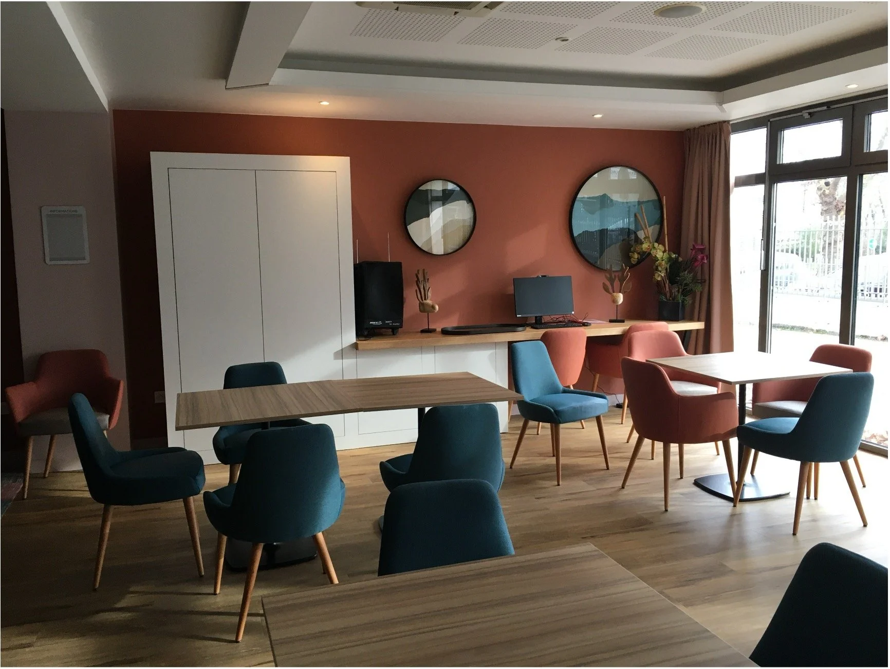 Salon avec des tables en bois des chaises bleus et rouges, des tableaux rond graphique, une armoire blanche et une baie vitrée.