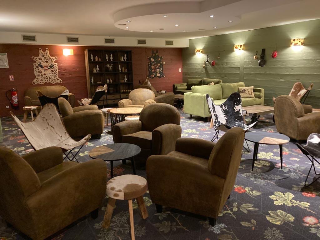 Salon composé de fauteuils club en cuir vieilli, de fauteuils en peau de vache, un canapé en velour vert, des tables basses en bois. Une bibliothèques en bois avec des objets décoratif, un mur en vert et rouge