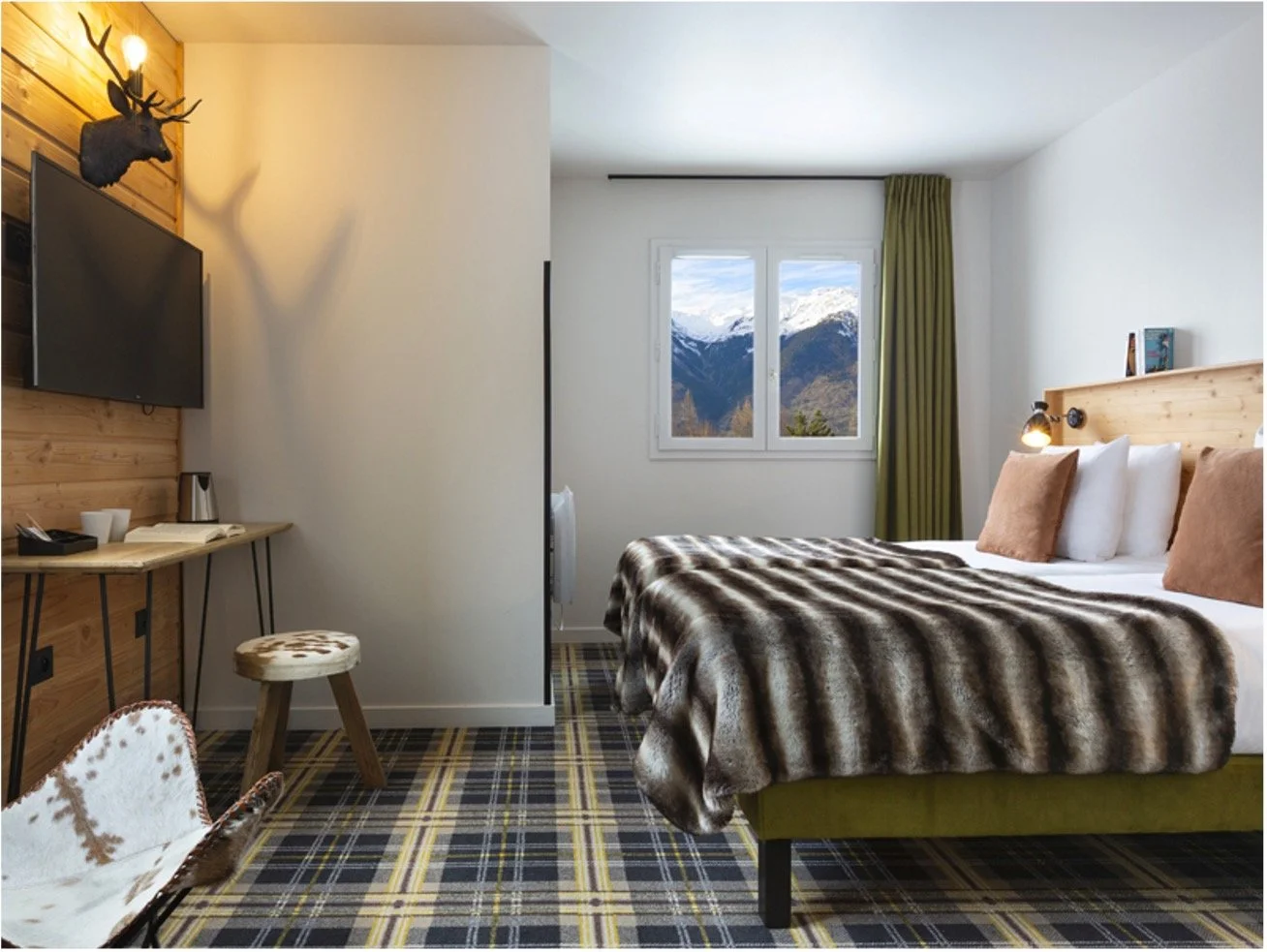 Chambre d'hotel à la montagne avec une télévison, un bureau, un mur en bois, un tabouret en peau de vache, un lit sur lequel est posé un plaid rayé noir et blanc et des coussins marron et blanc