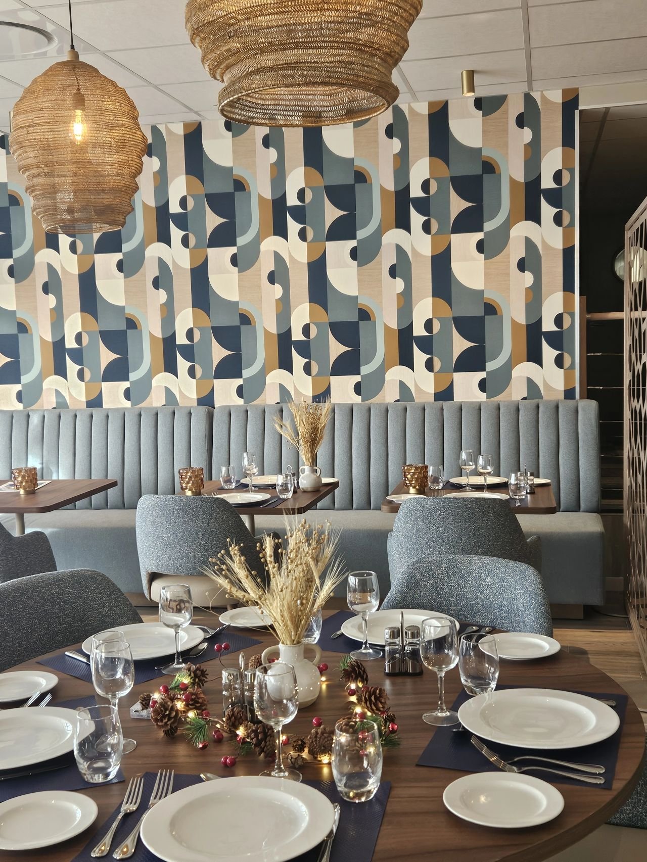 Salle à manger avec des tables dressées en bois, des chaises, des luminaires suspendus, une banquettes et un papier peint graphique bleu blanc et camel.