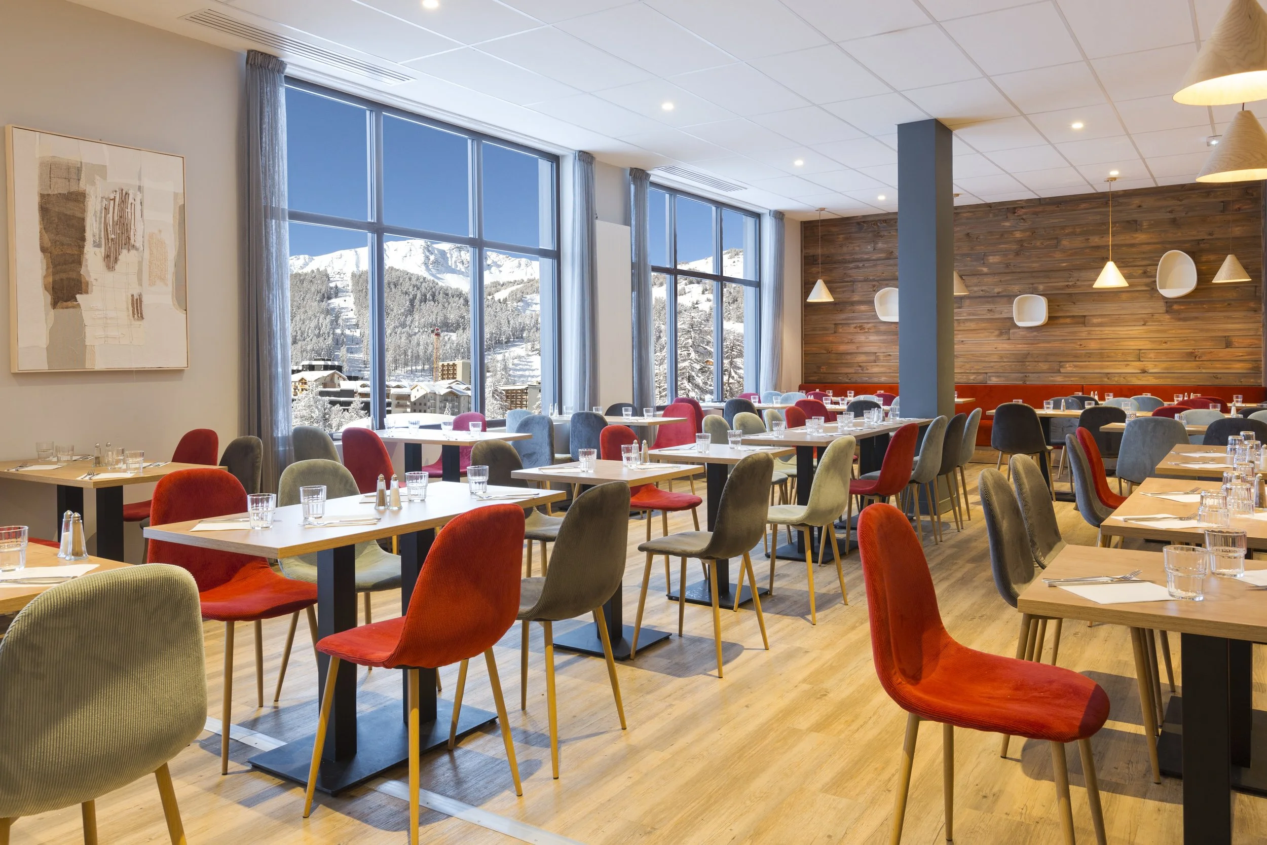 Salle à manger avec des table en bois, des chaises kaki et rouge, des lampe suspendues en forme de cône, des tableaux blancs, et deux baies vitrées qui donne la vue sur une montagne enneigée.