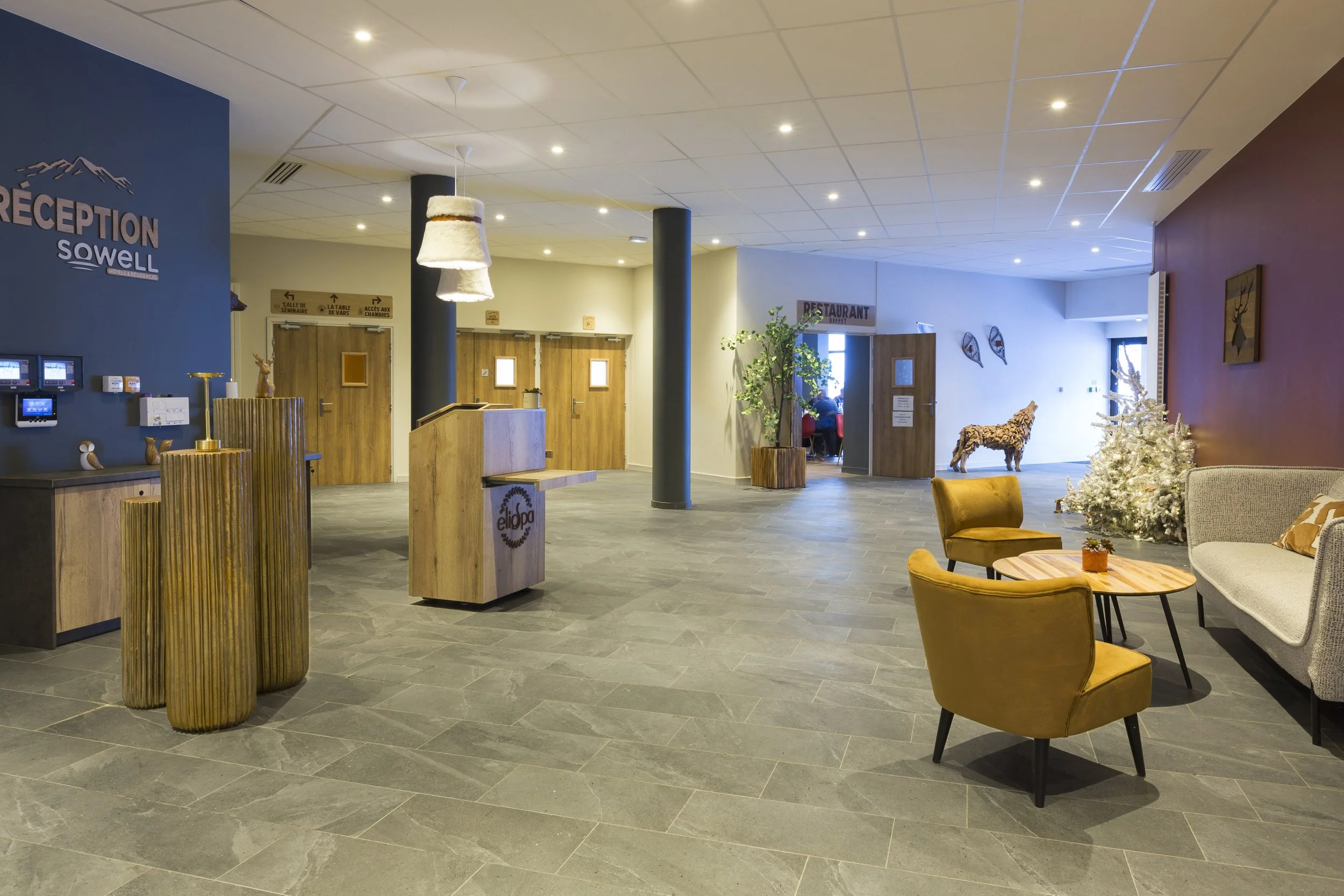 Hall d'accueil avec deux comptoir en bois, un mur et des piliers bleu marine, un sol avec un carrelage en pierre, des raquettes de ski accroché en mur, une table basse en bois deux canapé beige, une plante verte et une statue en bois en forme de loup