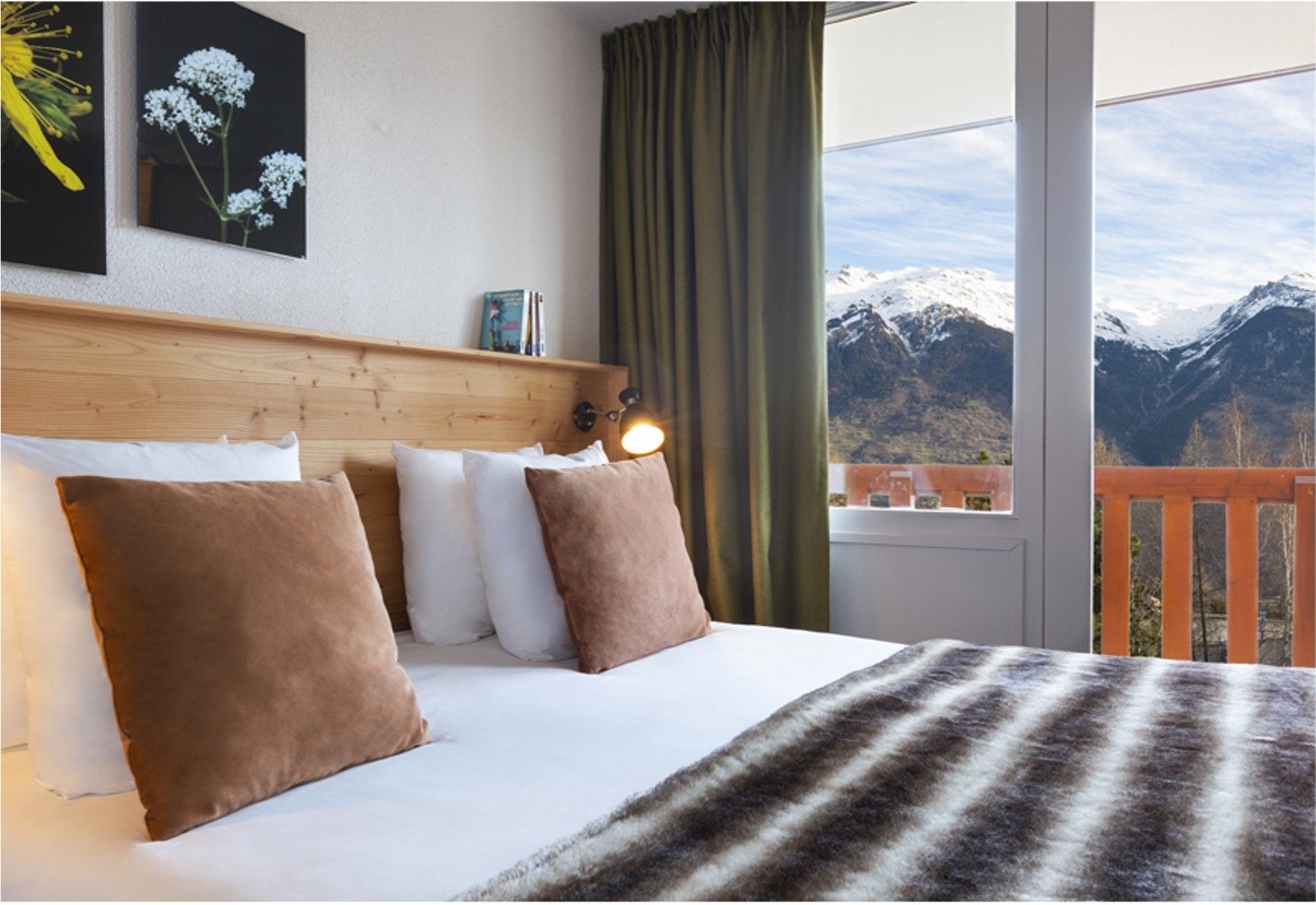 Chambre d'hôtel avec un lit sur lequel est posé des coussins blanc et marron ainsi qu'un plaid rayé noir et blanc. Deux tableaux de fleur et un rideaux vert. Fenêtre ou l'on voit à travers la montagne
