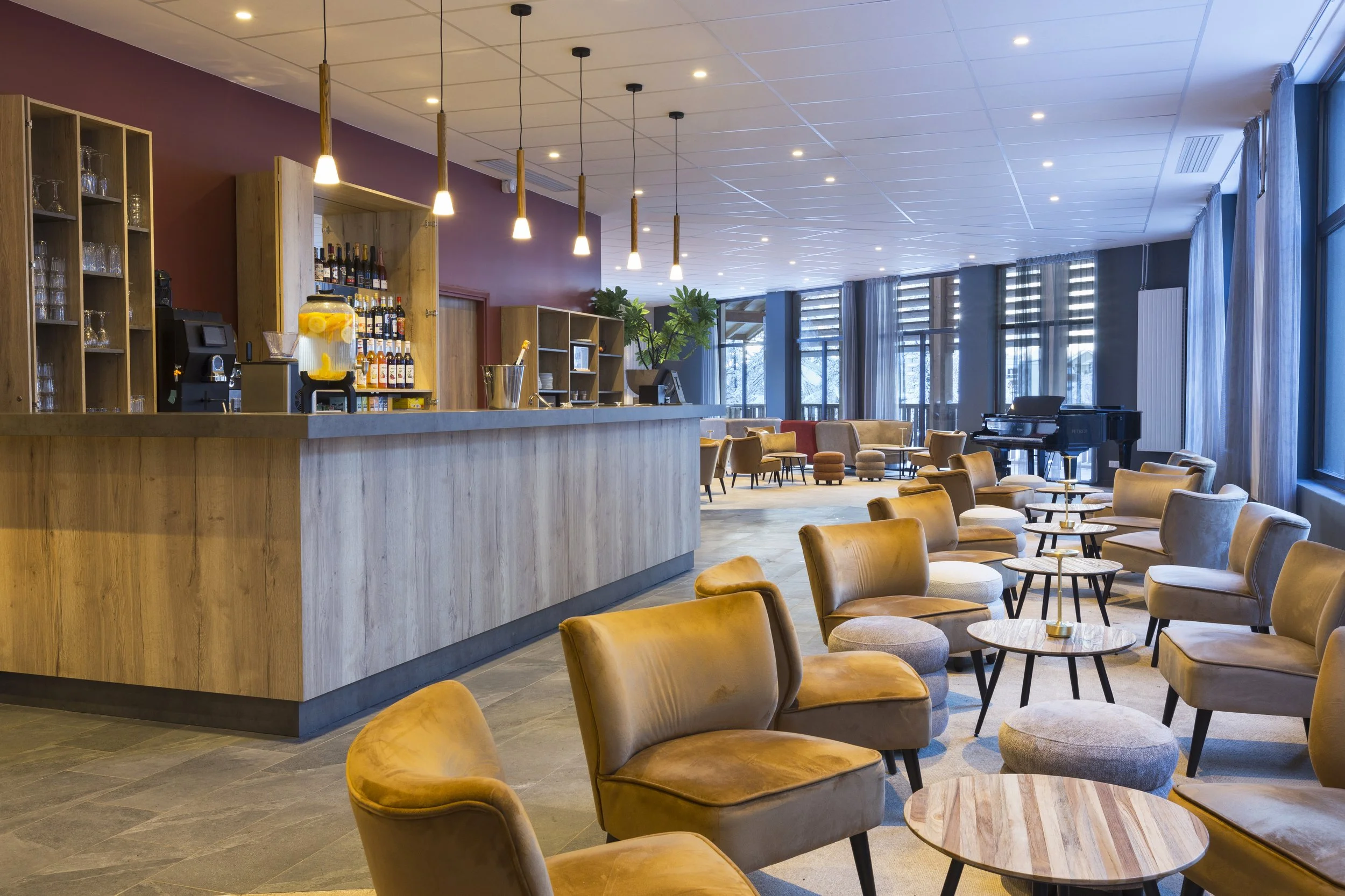 Bar d'hôtel avec un comptoir en bois, des luminaires suspendues, des chaises basses beige et des tables basses en bois.