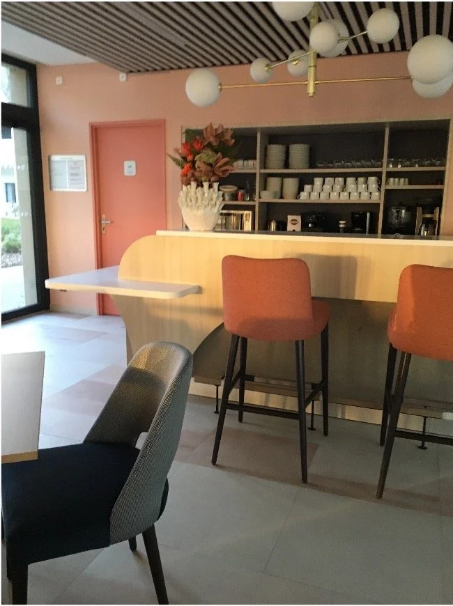 Comptoir de bar sur lequel est posé un bouquet de fleur dans un vase en forme de corail avec des chaises rose saumon et bleu. Une suspension composé d'une dizaine de globe blancainsi qu'un arrière comptoir avec sa machine à café, verre, tasse