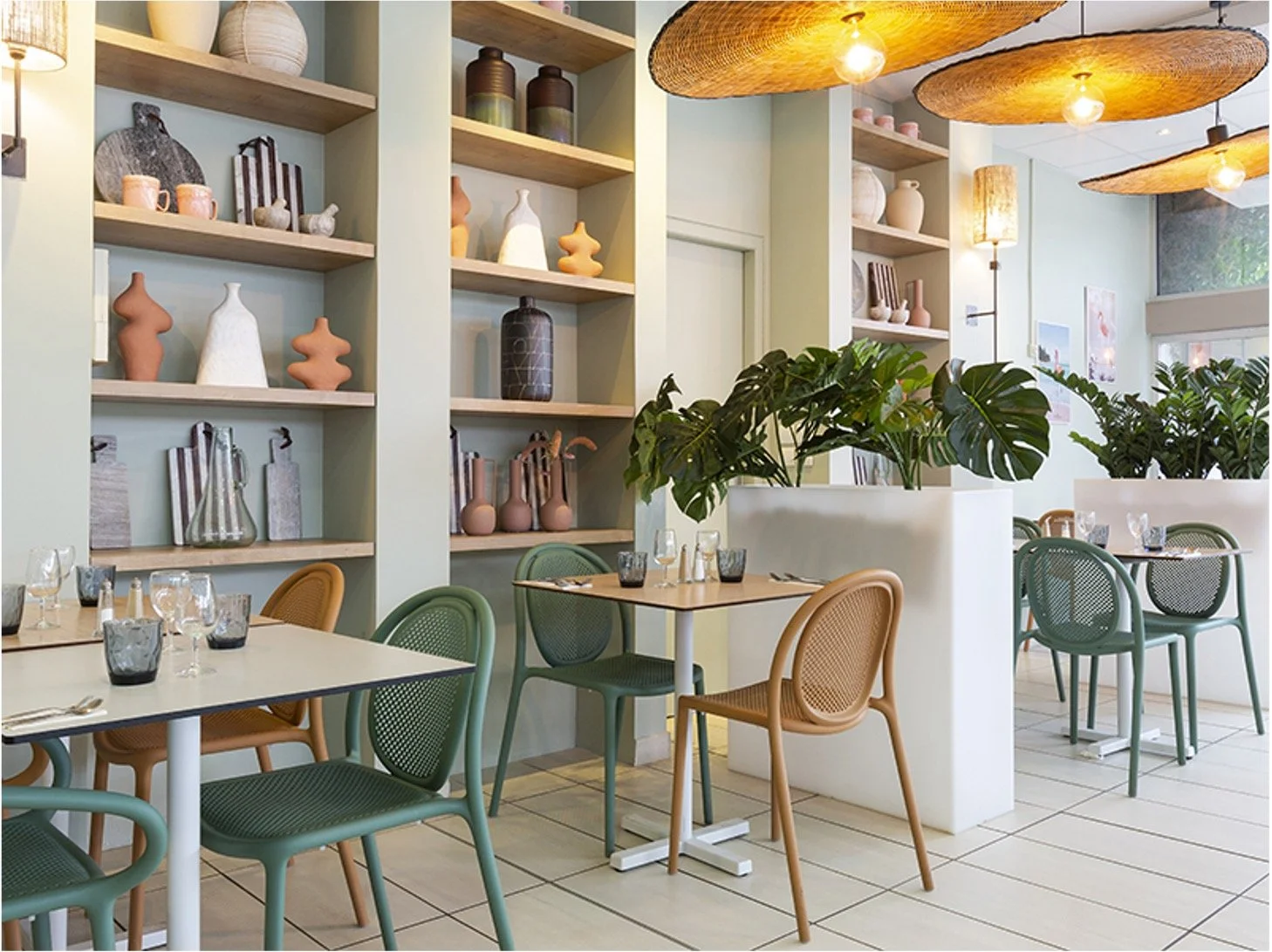 salle de restaurant avec des tables, des chaises vertes et terracotta. Séparation avec plante verte. Etagère avec objets décoratifs à l'intérieur style bord de mer. Suspension en forme de chapeau de paille camarguais.