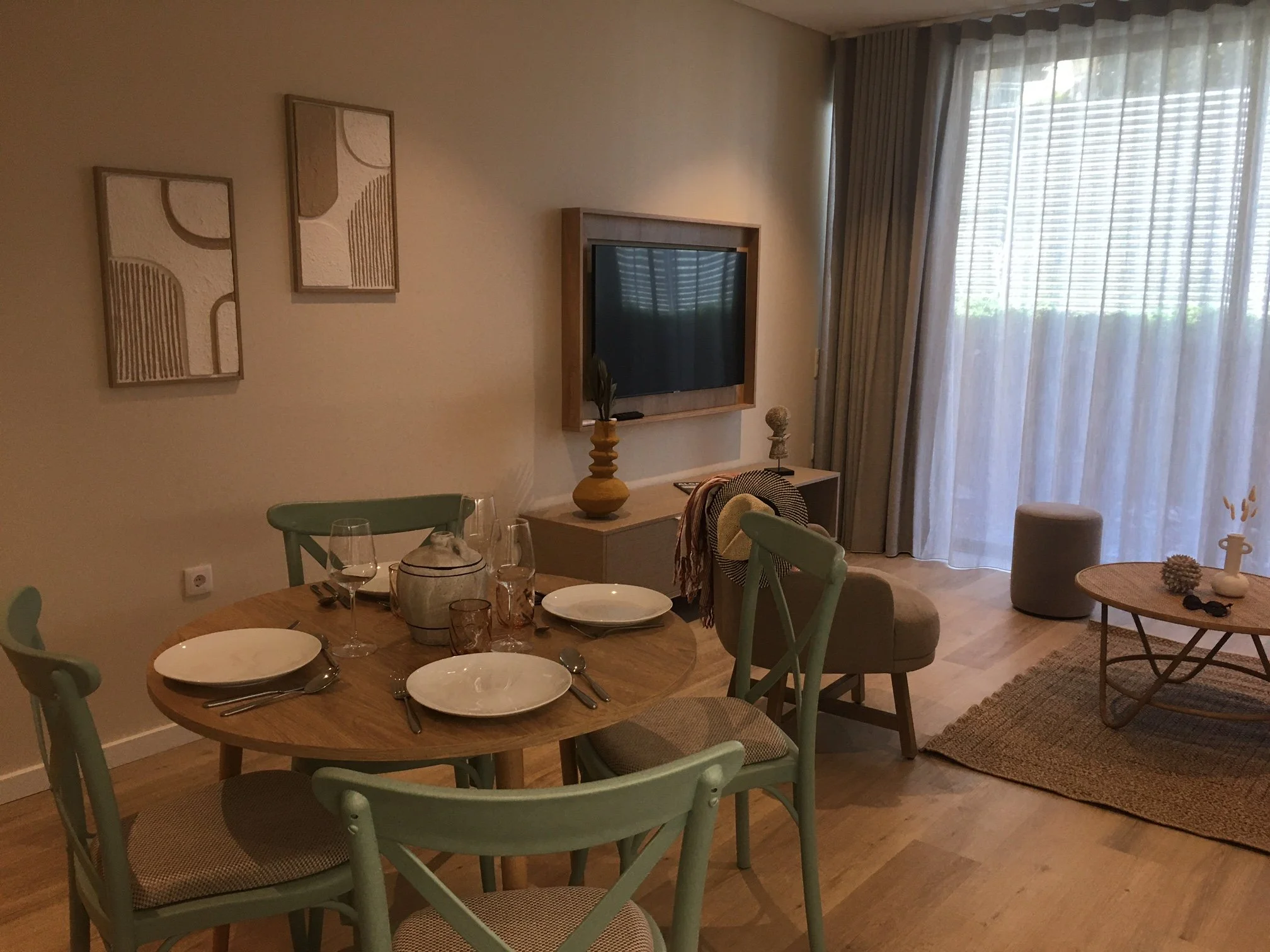 Salon avec 4 chaises vertes, une table en bois, des assiettes, des verres, deux tableaux, une télévision, une commode, un tapis, des rideaux, un tabouret et un fauteuil.