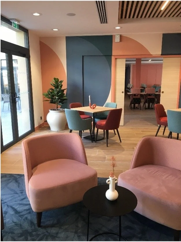Salon avec des fauteuils rose saumon, des chaise rouge t bleu avec une plante dans une jarre ainsi qu'un papier peint géométrique terracota et bleu