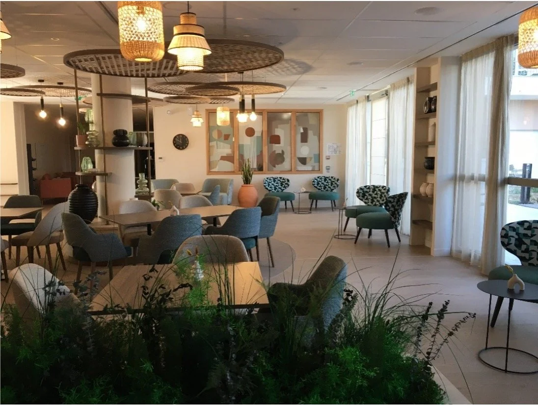 Salon de bar composé d'une jardinière avec des plantes verte, des tables en bois, des chaises et fauteuils bleu, des objets décoratif sur des étagère, des tableaux avec des formes géométrique ainsi que des luminaires suspendu au plafond