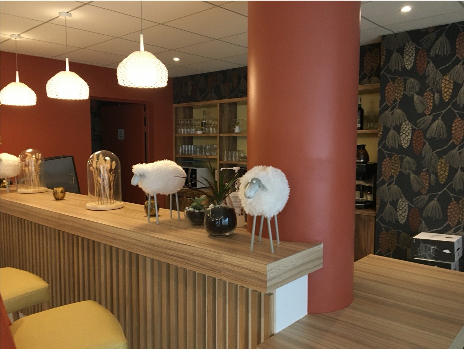 Comptoir de bar en bois avec des chaises haute, un pilier rouge, un papier peint illustrant des pommes de pins, des objets décoratifs.