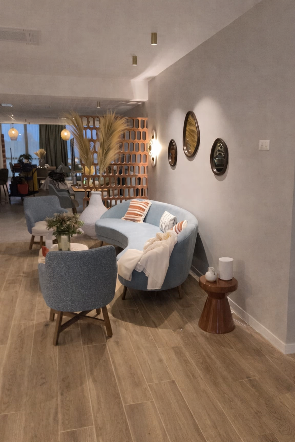 Hall d'accueil avec un claustra de brique en terre cuite, un canapé bleu ciel avec des coussins dessus, deux fauteuils, des tables basses. Trois miroirs accrochés sur le mur. Herbe de la pampa et un claustra en brique au fond.