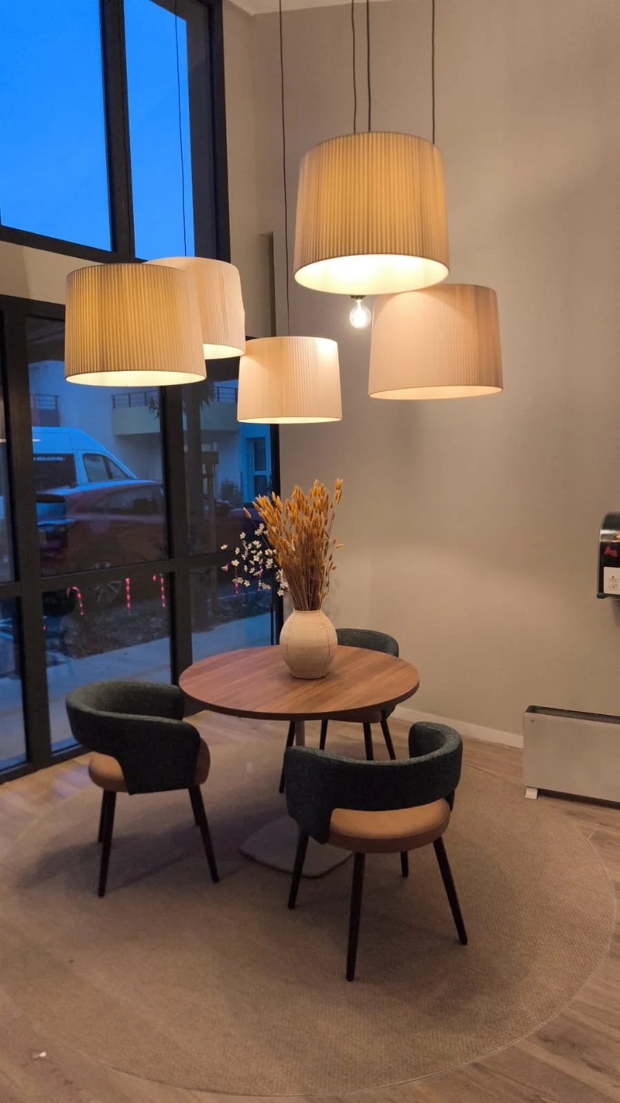 Hall d'accueil avec une table en bois, des chaises, un bouquet de fleur dans un vase, une baie vitrée et des luminaires suspendus au plafond.