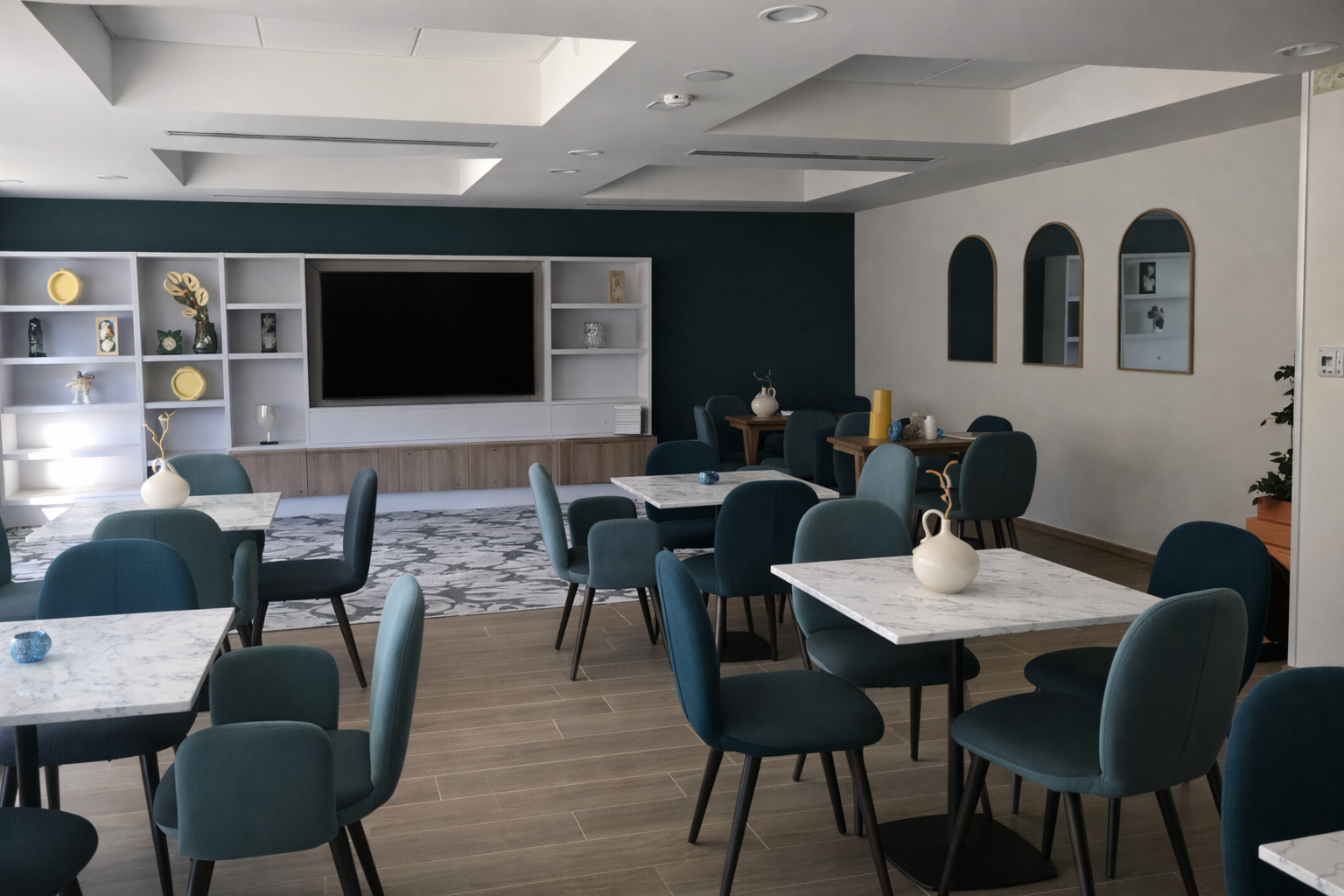 Salle à manger avec des tables en marbre, des chaises bleus, une télévision, étagère avec des objets décoratifs à l'intérieur, un mur bleu. Trois miroirs posé sur un mur blanc.