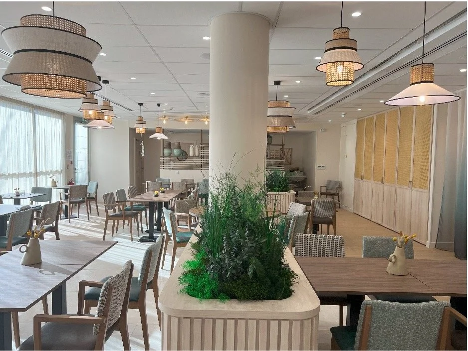 Salle à manger composé d'une jardinière, des tables en bois, des chaises bleu gris, des luminaires suspendu au plafond et un pilier au centre