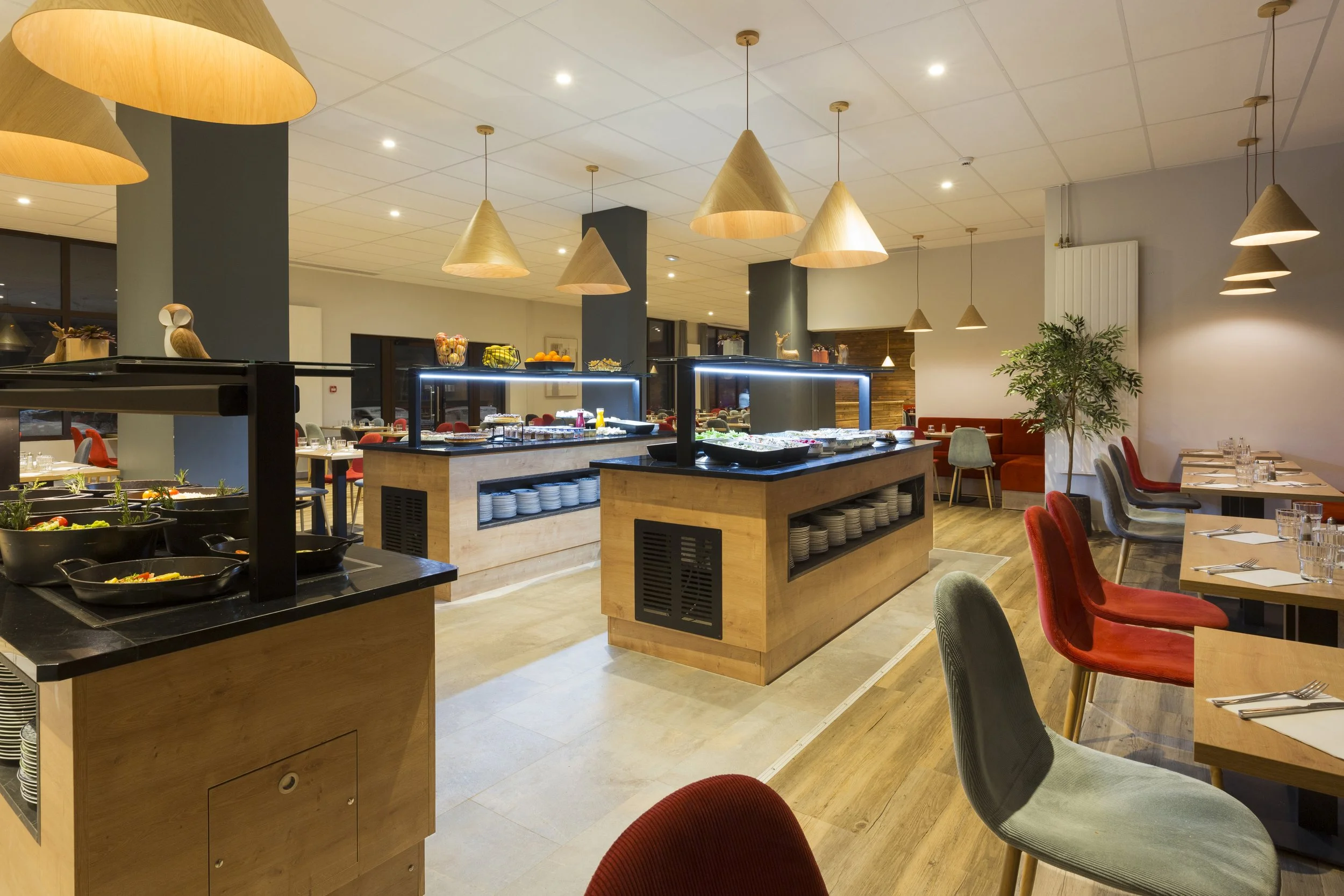 Buffet d'hôtel en bois avec des lampes suspendues en forme de cône, des chaises grises et rouge avec des piliers bleu marine.