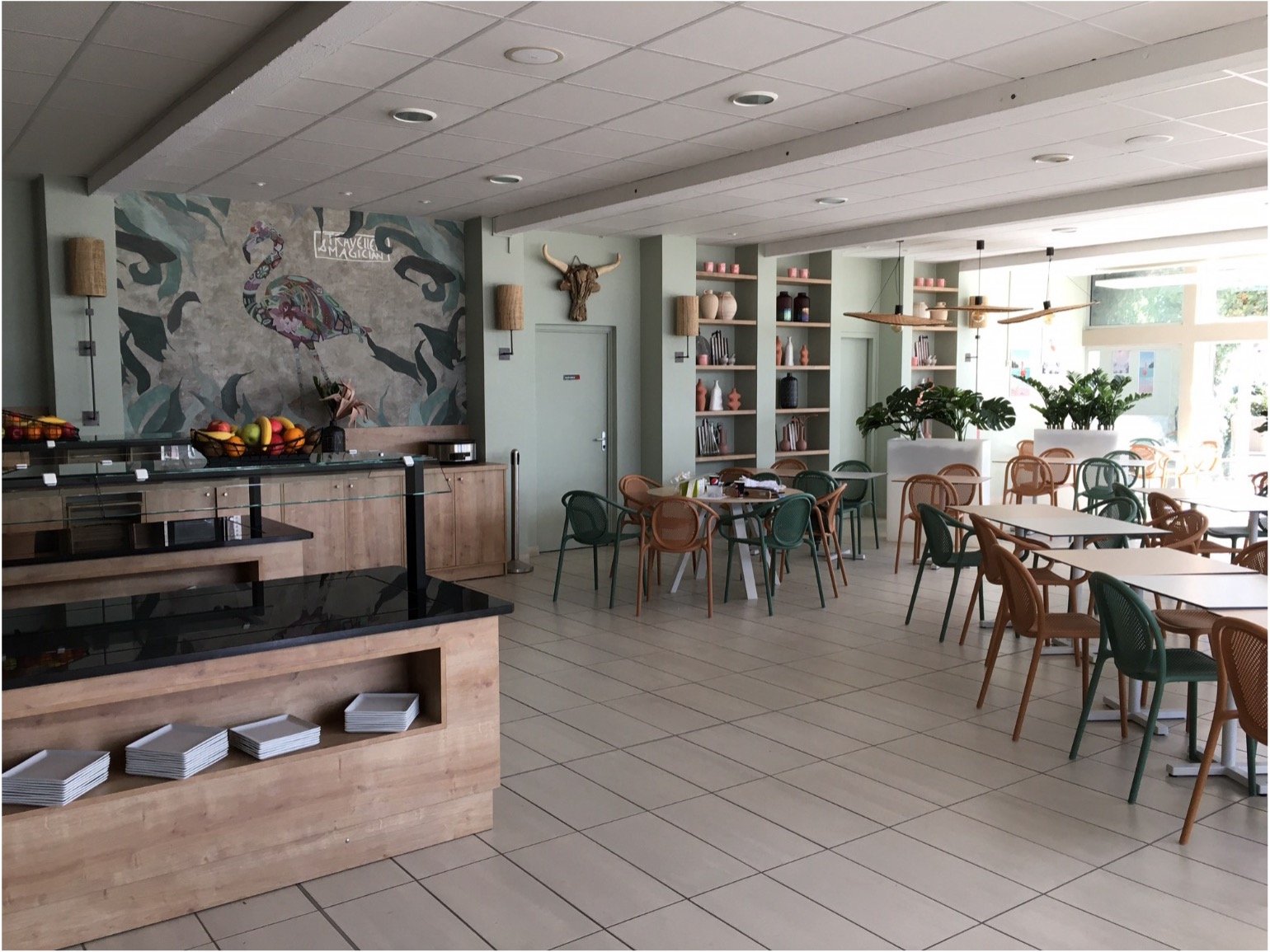 Salle de restaurant avec un buffet desserte, un papier peint illustrant un flamant rose au dessus. Des tables en bois avec des chaises verte et terracota. Des appliques, une tete de taureau et une porte verte. Une étagère avec des objets décoratifs.