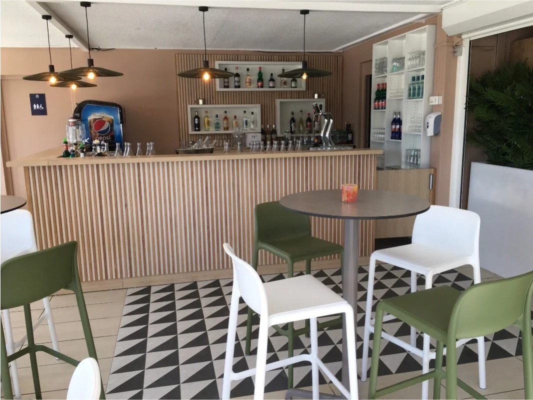 Comptoir de bar avec des luminaires suspendus au plafond, un sol graphique noir et blanc et tabourets haut blanc et vert.