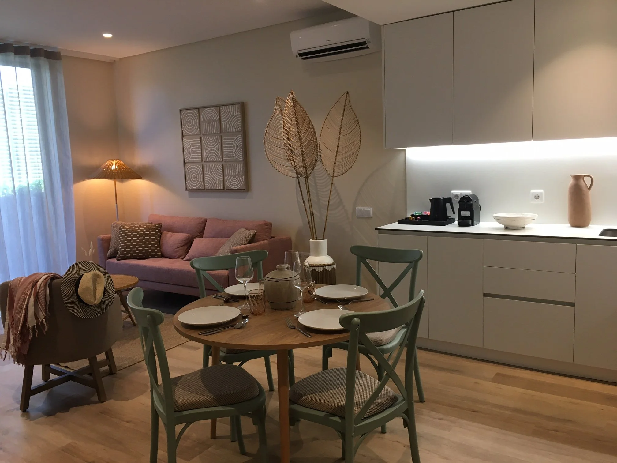 Salon avec quatre chaises vertes, des placards blancs, un tableau, une lampe, une cafetière, un vase, des assiettes, des verres, un canapé rouge et un fauteuil avec un chapeau, de grande feuille en bambou dans un vase.