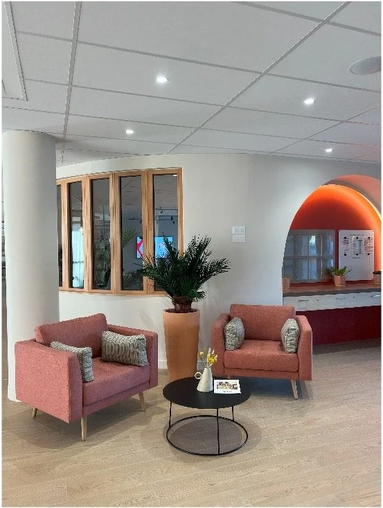 Hall d'accueil avec des fauteuils rose saumon, une jarre avec une plante à l'intérieur, un pilier blanc et des fenêtre.Un comptoir d'accueil sur un fond rouge.