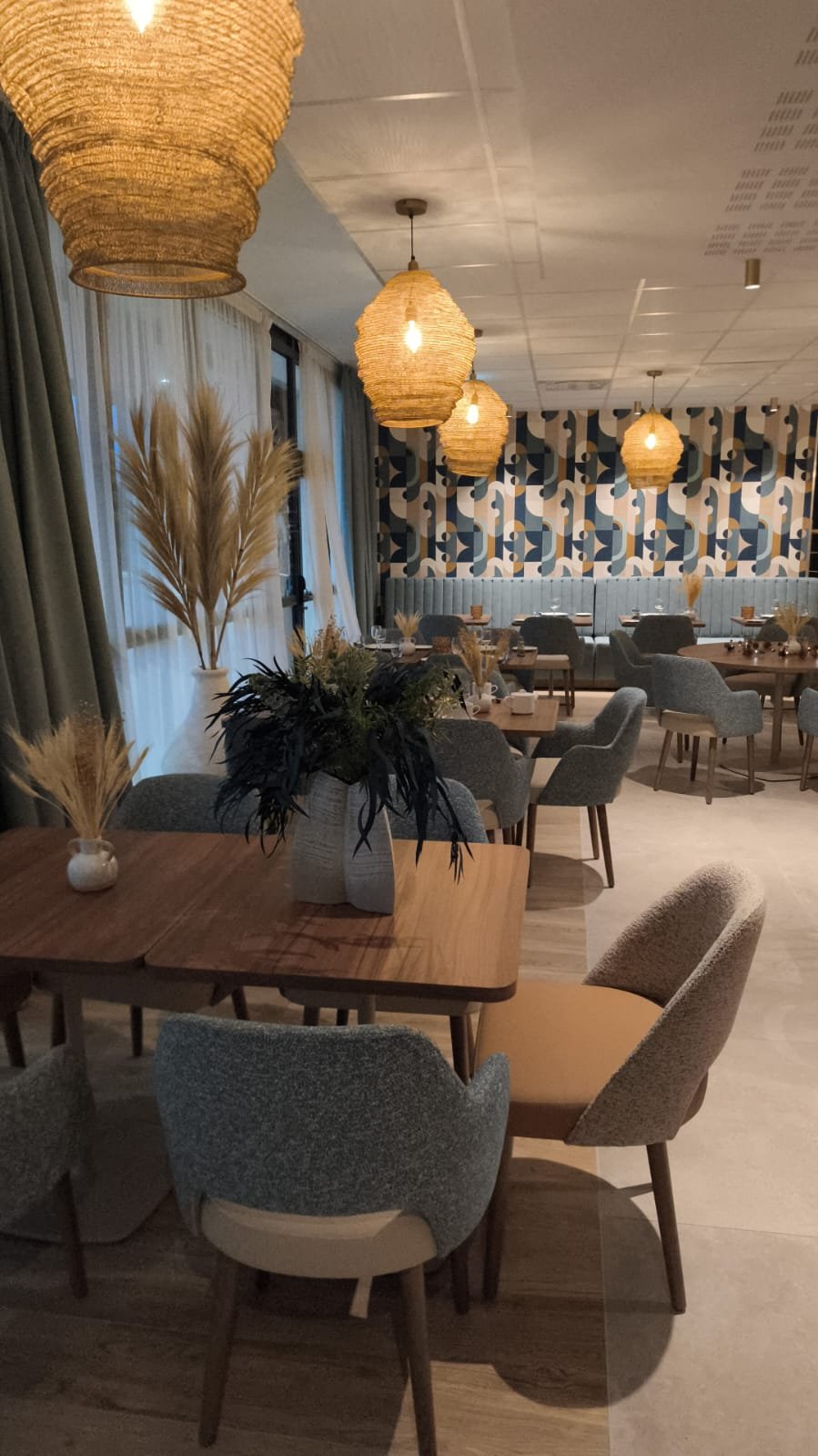 salle à manger avec des tables en bois, des chaises blanche et bleu, des bouquets de fleur, des luminaires suspendus en plafond, des herbes de la pampa dans un vase, une banquette bleu et un papier peint couleur bleu, blanc et camel.