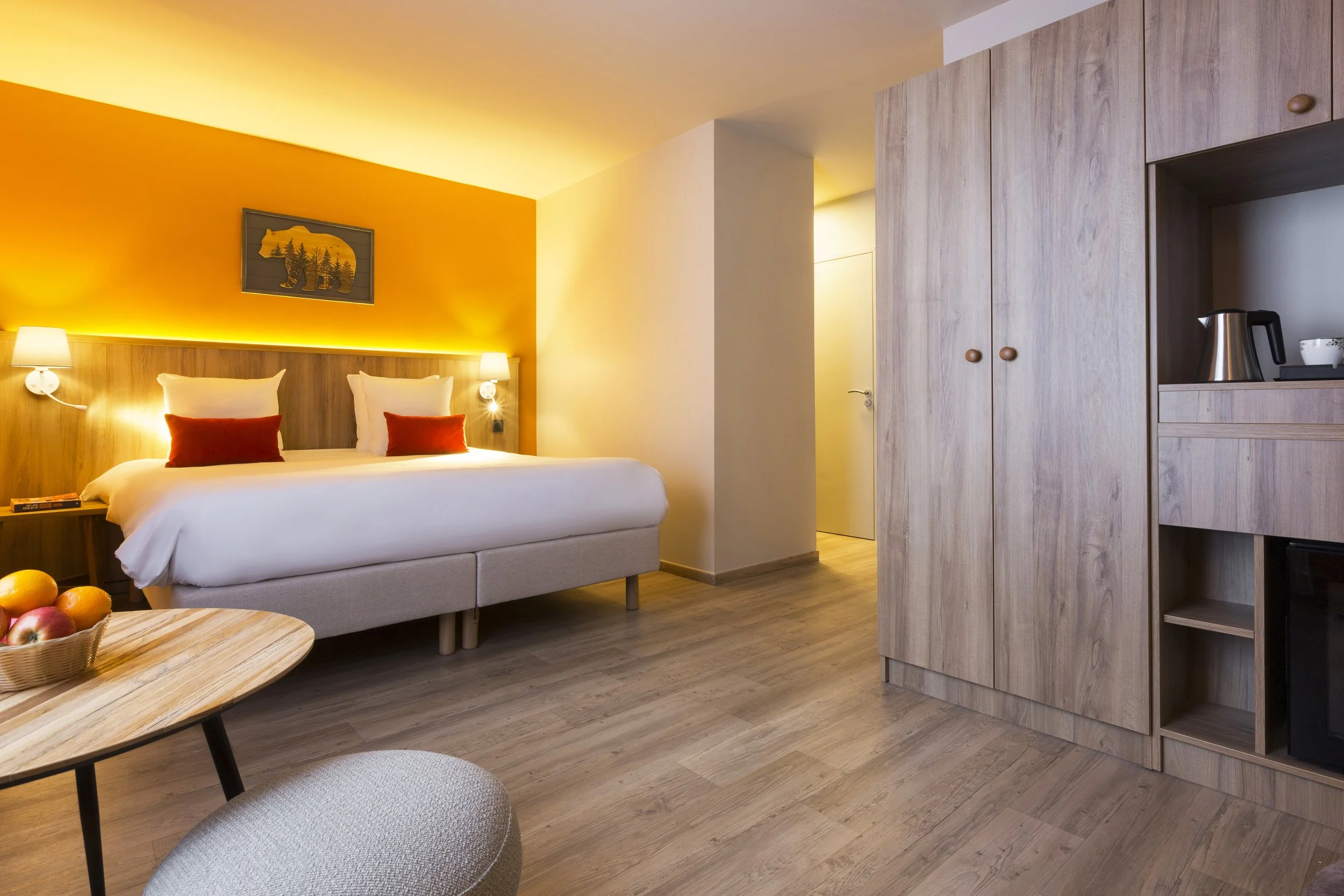 Chambre d'hôtel avec un lit, des coussins blanc et rouge, un tableau qui représente un ours, une armoire en bois, un mur peint en jaune, une tête de lit en bois, deux lampe de séjour et une table basse en bois.