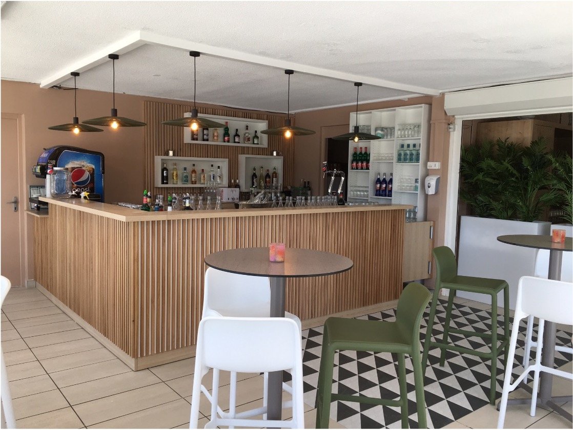 Comptoir de bar avec des luminaires suspendus au plafond, un sol graphique noir et blanc et tabourets haut blanc et vert. Des Etagères avec des bouteilles posées dessus.