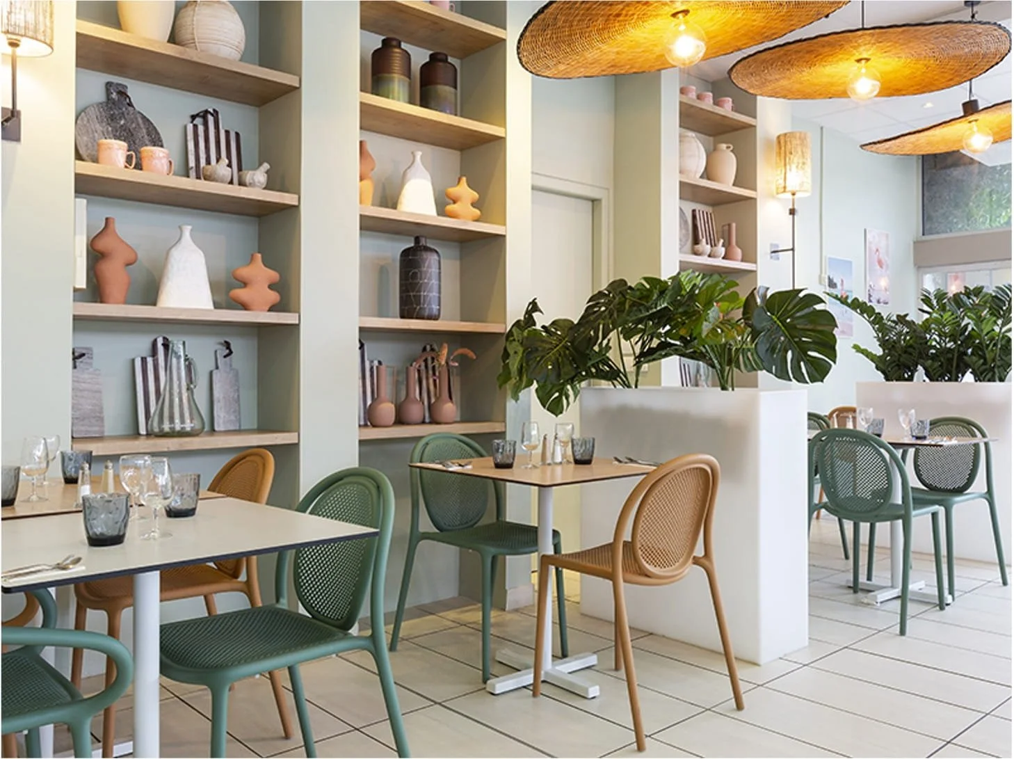 salle de restaurant avec des tables, des chaises vertes et terracotta. Séparation avec plante verte. Etagère avec objets décoratifs à l'intérieur style bord de mer. Suspension en forme de chapeau de paille camarguais.