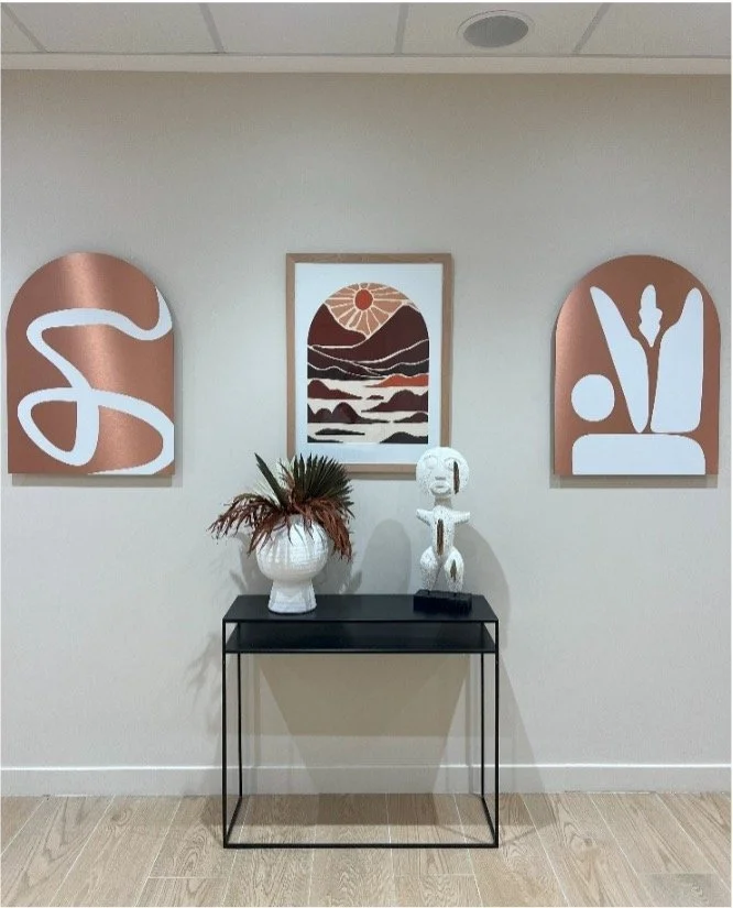 Console noire sur laquelle est posé un vase blanc avec des pleure et une statue en bois mexicaine blanche, avec trois tableaux dont deux en metal cuivré sur un mur blanc