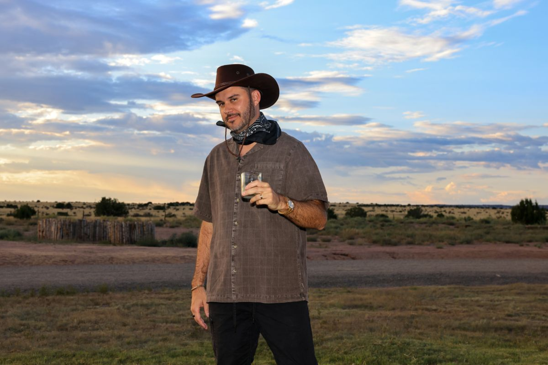 A man dressed in cowboy attire holding a glass in a rural outdoor setting during sunset, with a cloudy sky and open fields in the background.