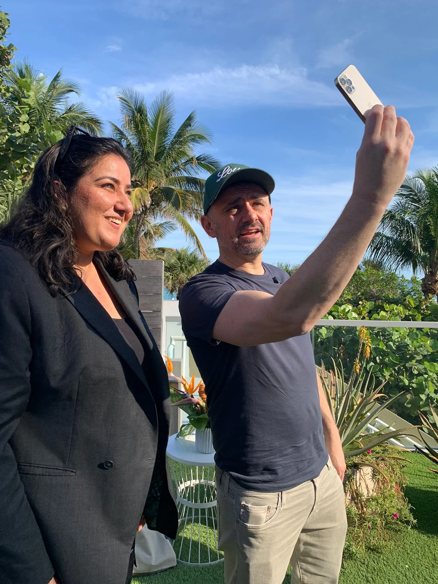 A man and a woman are standing outdoors on a sunny day, taking a selfie. The woman is smiling, and the man is looking at the phone. There are palm trees and plants in the background.