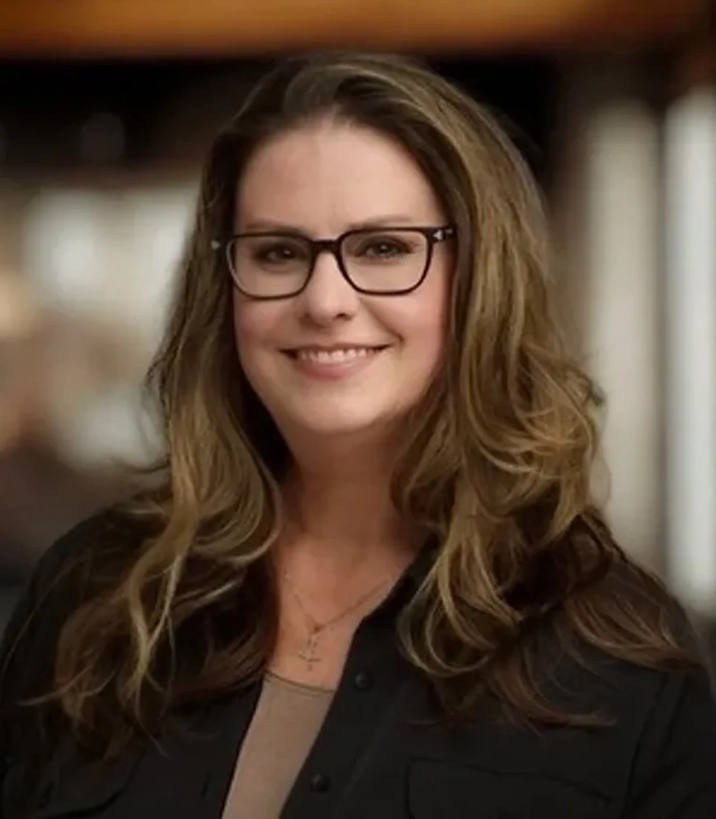 A woman with long, wavy brown hair and glasses smiling in an indoor setting.