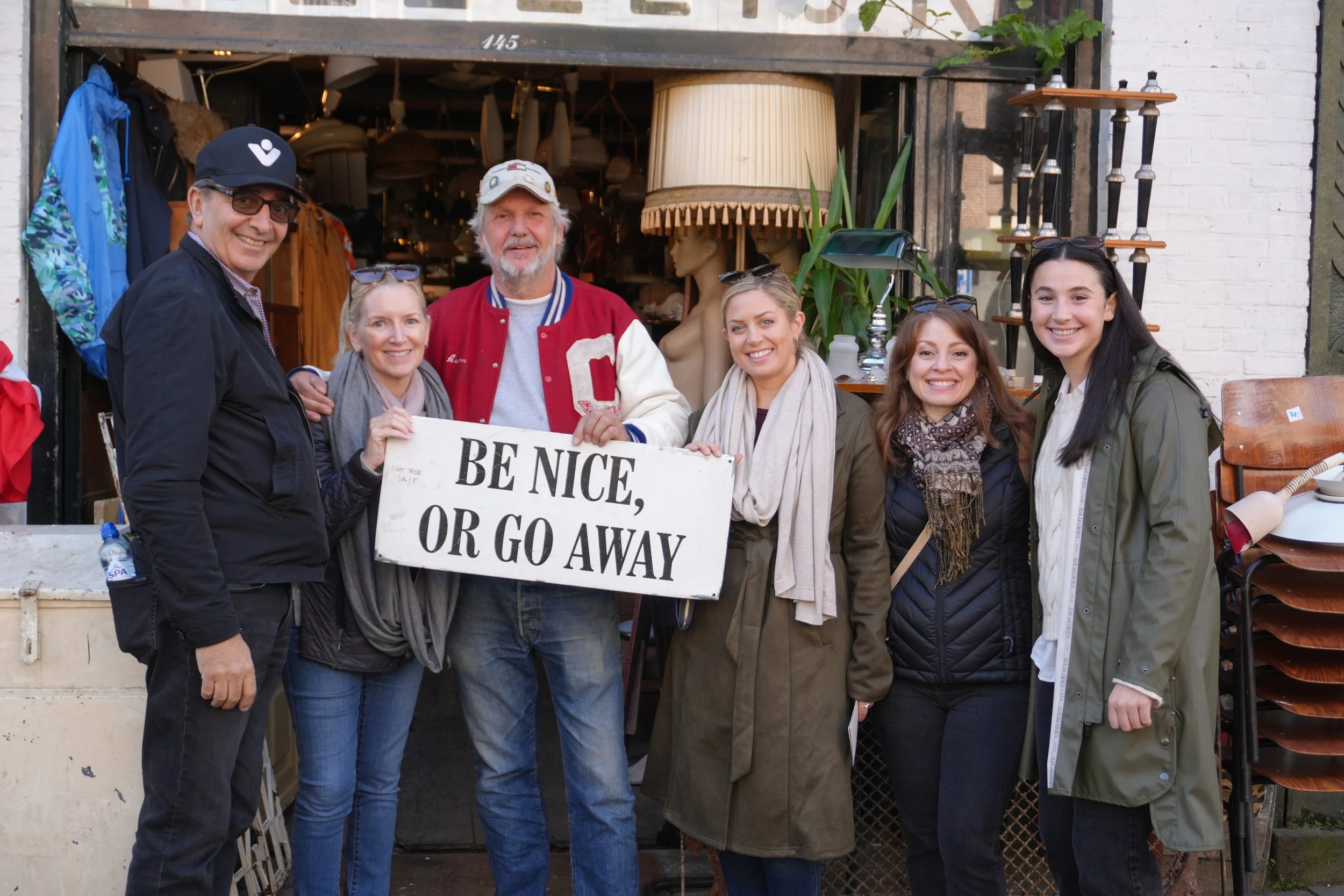 Group of seven people standing outside a shop, one man holding a sign that reads "Be nice, or go away". They are smiling and dressed casually, with various items visible inside the shop behind them.