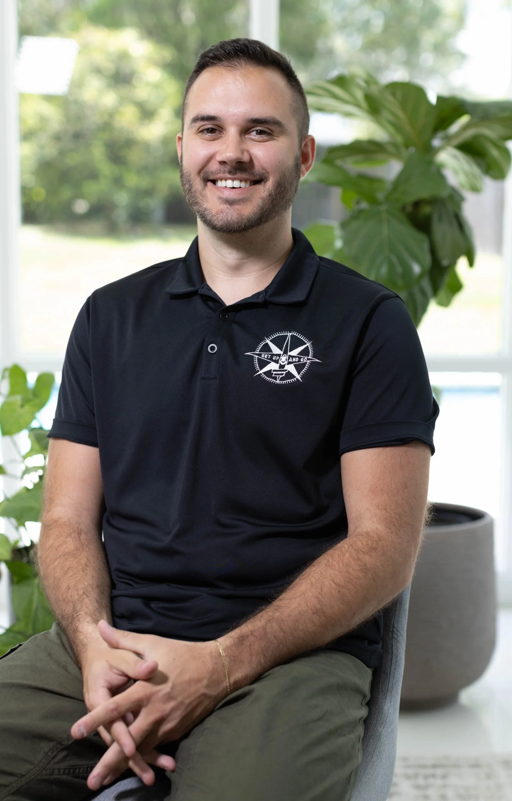 A young man with short dark hair and a beard, smiling, sitting indoors in front of a large window with trees outside. He is wearing a black polo shirt with a white logo on the left chest and khaki pants.