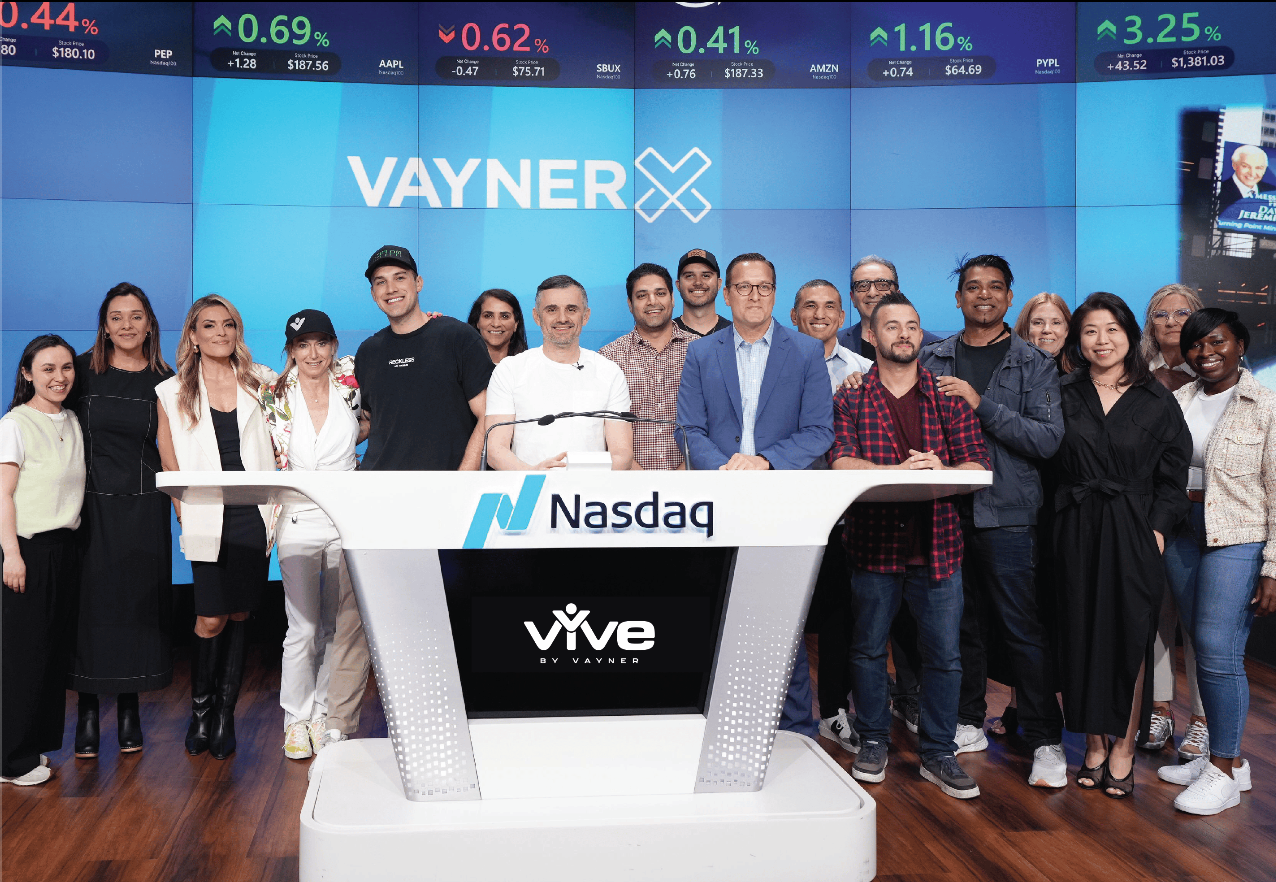 Group of diverse people standing behind a Nasdaq podium during a corporate event or presentation. Large digital stock market display in the background showing stock price changes.