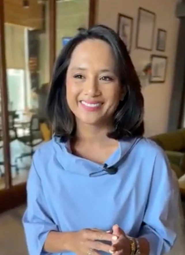 A woman with shoulder-length black hair smiling, wearing a light blue blouse, in an indoor setting with framed pictures on the wall behind her.
