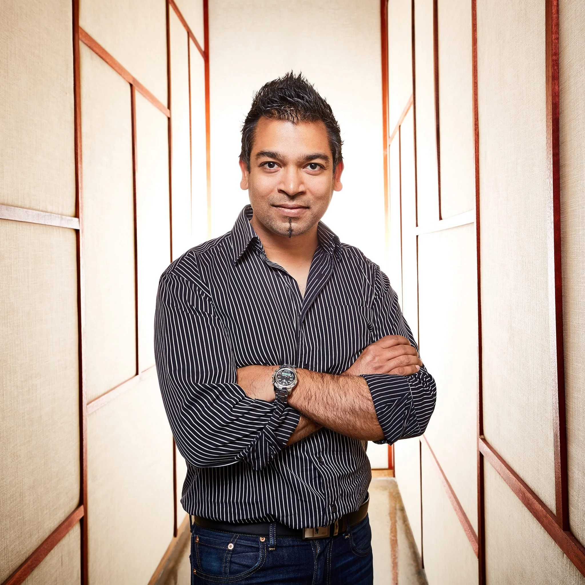 A man with crossed arms standing in a hallway with beige wall panels and wood trim, backlit by bright light.