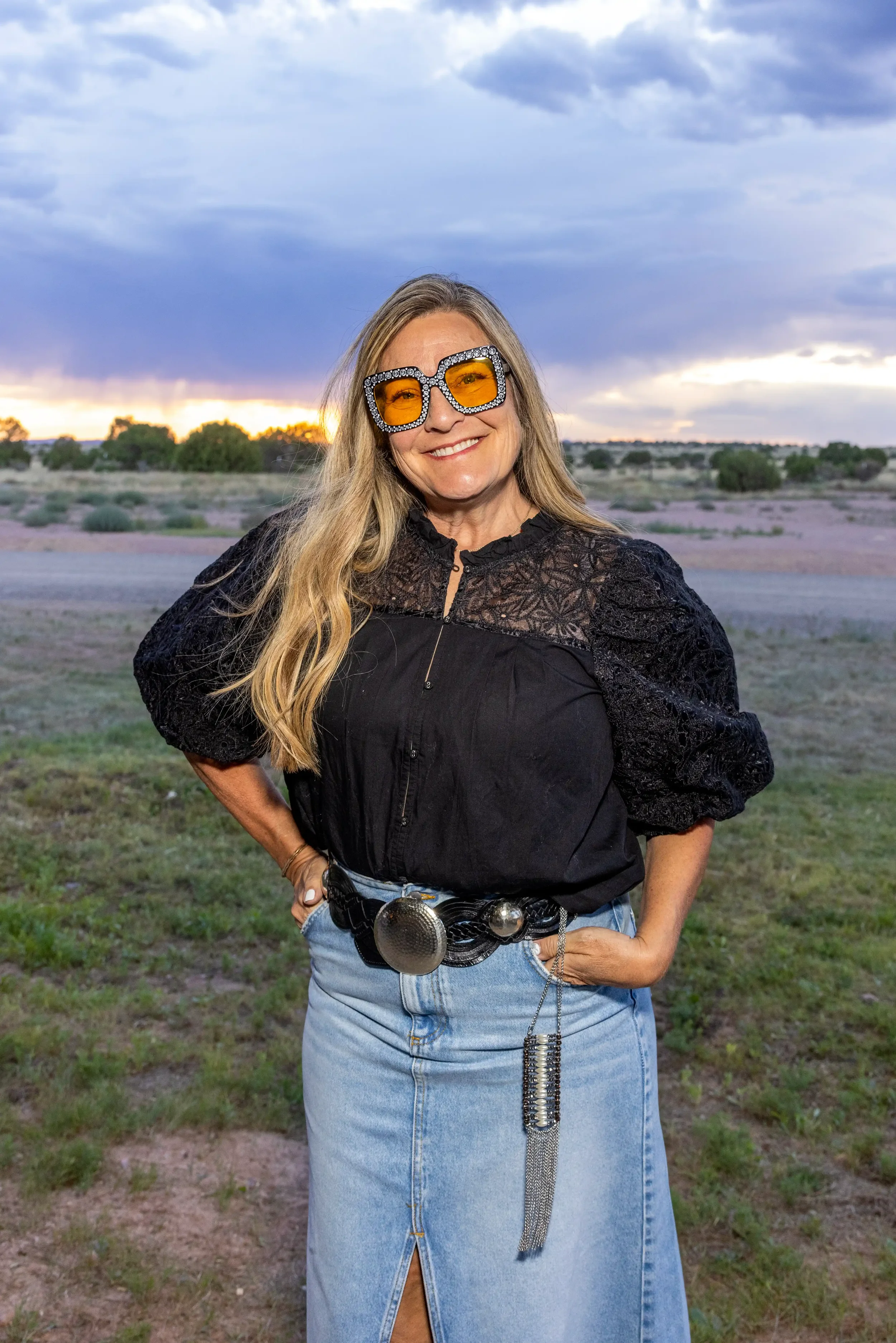 A woman wearing unique sunglasses with orange lenses and rhinestones, a black lace-detail blouse, and a denim skirt with a large belt and decorative chain standing outdoors during sunset with a cloudy sky and green landscape in the background.