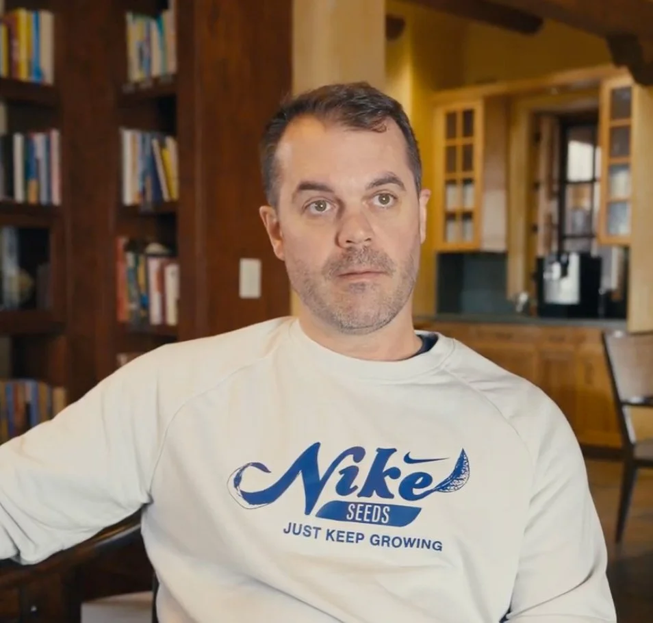 A man wearing a white sweatshirt with a seed company's logo sitting in a cozy wooden kitchen or living room.