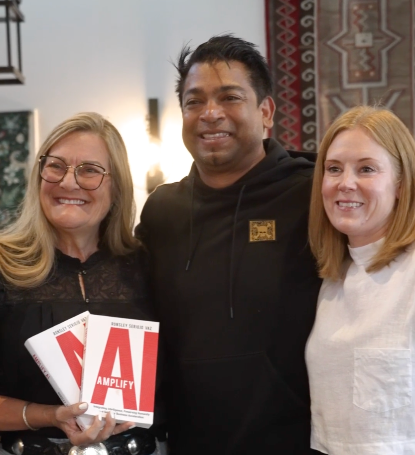Three people smiling and posing for a photo, two women and one man, with one woman holding books titled 'Amplify'.