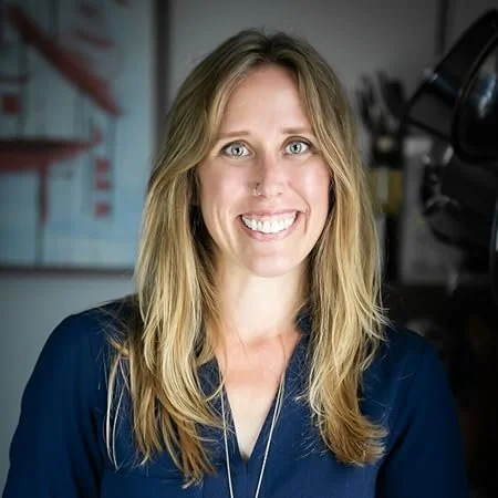 A smiling woman with long blonde hair, wearing a dark blue top, in an indoor setting with a blurred background.