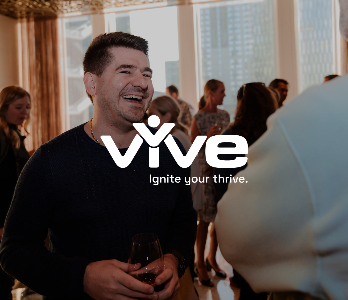 Man smiling and talking at a social gathering with other people in the background, holding a glass of wine, in a well-lit room with large windows.