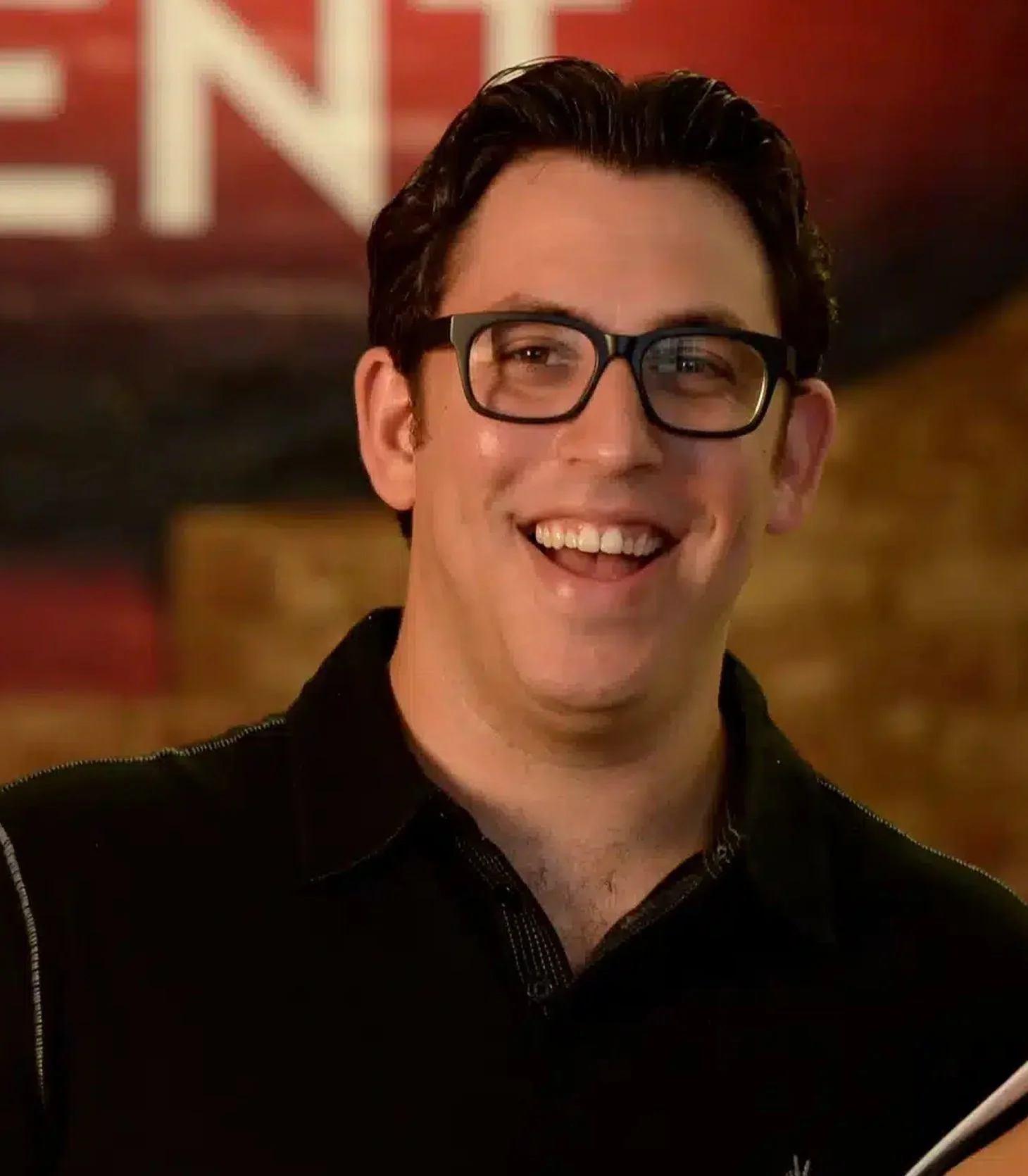 A smiling man with black hair, wearing glasses, in a dark shirt with a background featuring warm lighting and some text