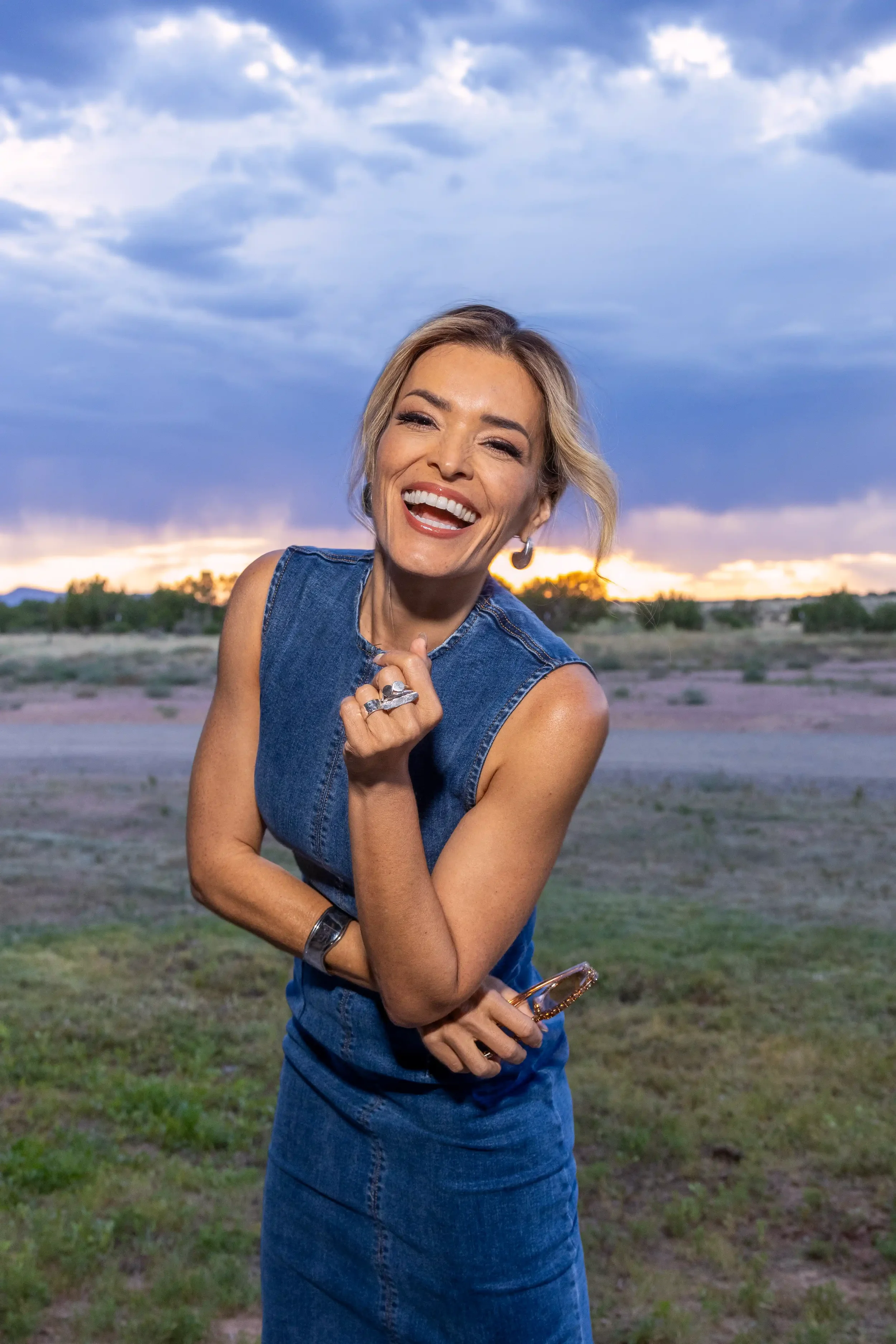 A smiling woman with light hair styled in loose waves, wearing a sleeveless denim top and skirt, holding sunglasses, standing outdoors at sunset with a partly cloudy sky and desert landscape in the background.