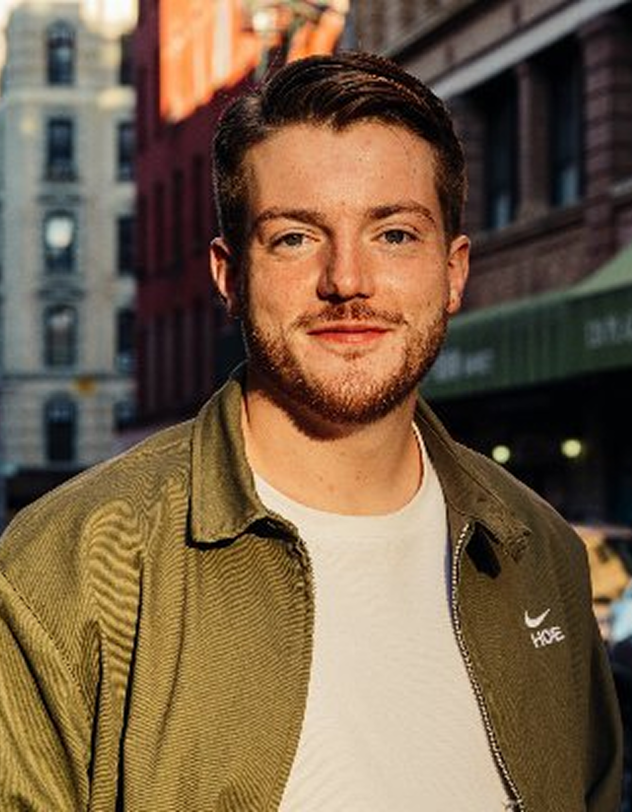 A young man with light skin, short dark hair, and a beard, standing outdoors in an urban setting with brick buildings, wearing a green Nike jacket and white shirt, smiling at the camera.