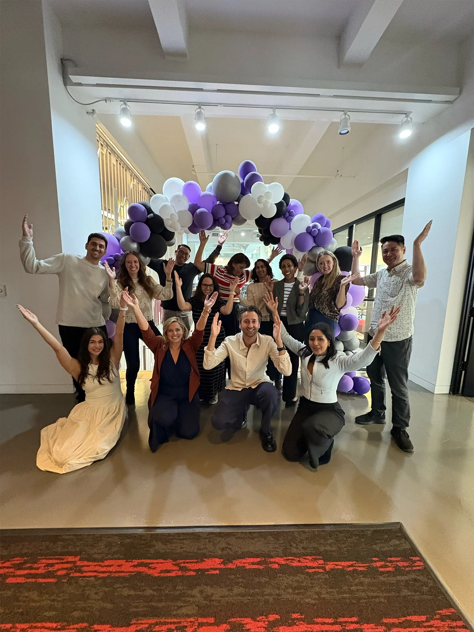 A group of people celebrating inside with purple, black, white, and gray balloon arch.