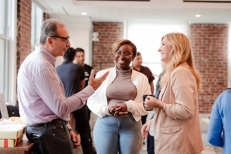 A diverse group of people sitting around a table during a meeting or workshop. One woman is standing and talking, while others sit and listen. The table has papers, markers, stickers, glasses, and drinks on it.