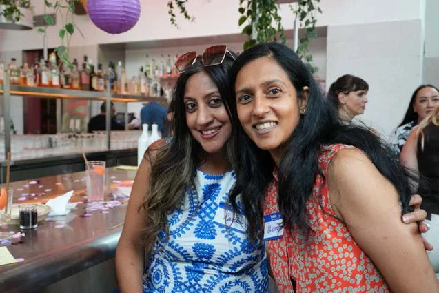 Two women smiling at a social event, with a bar and other people in the background.