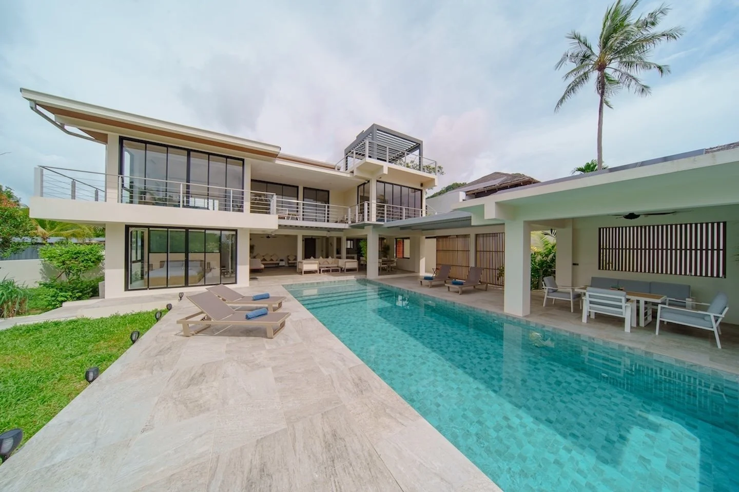 Modern private villa  with large outdoor swimming poo and roof top with sea viewsl, lounge chairs, and shaded patio area, featuring multiple balconies and large glass windows, set amid lush greenery and a tropical tree.